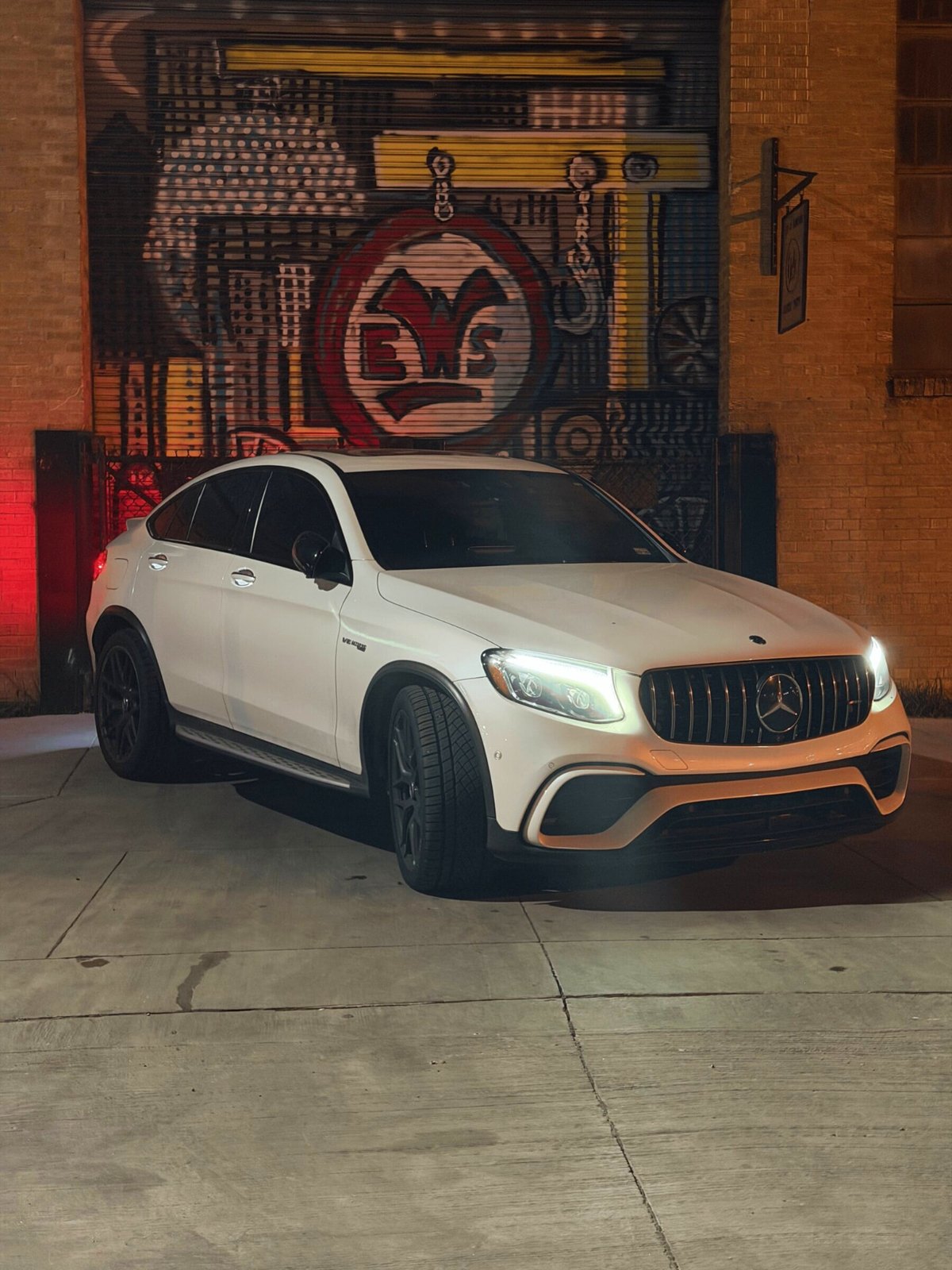 Mercedes-AMG GLC 63 S front three-quarter with headlights on against a painted mural wall at night